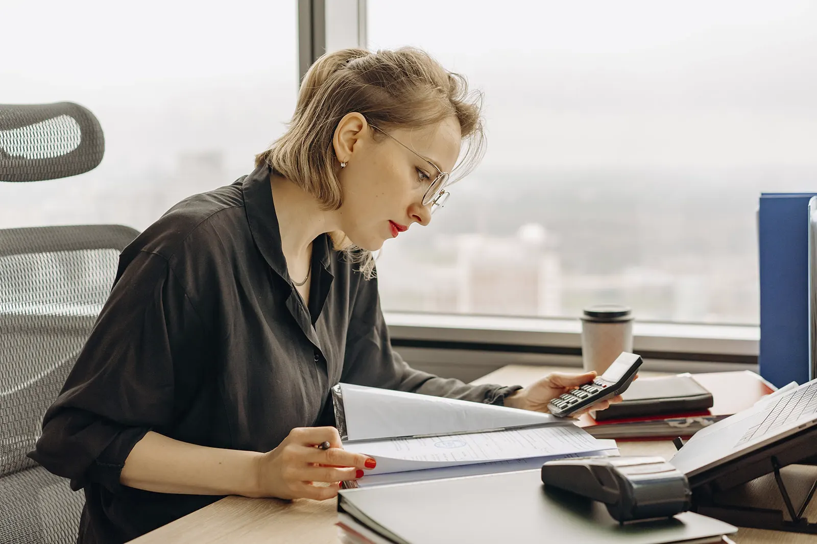 Mujer con gafas revisando documentos y usando una calculadora en un escritorio de oficina